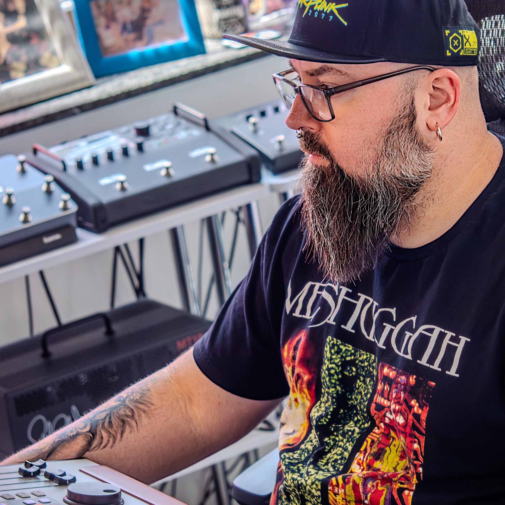 Man with a beard and glasses wearing a black t-shirt with a graphic design, sitting in front of electronic equipment.