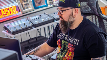 Man with a beard and glasses wearing a black t-shirt with a graphic design, sitting in front of electronic equipment.